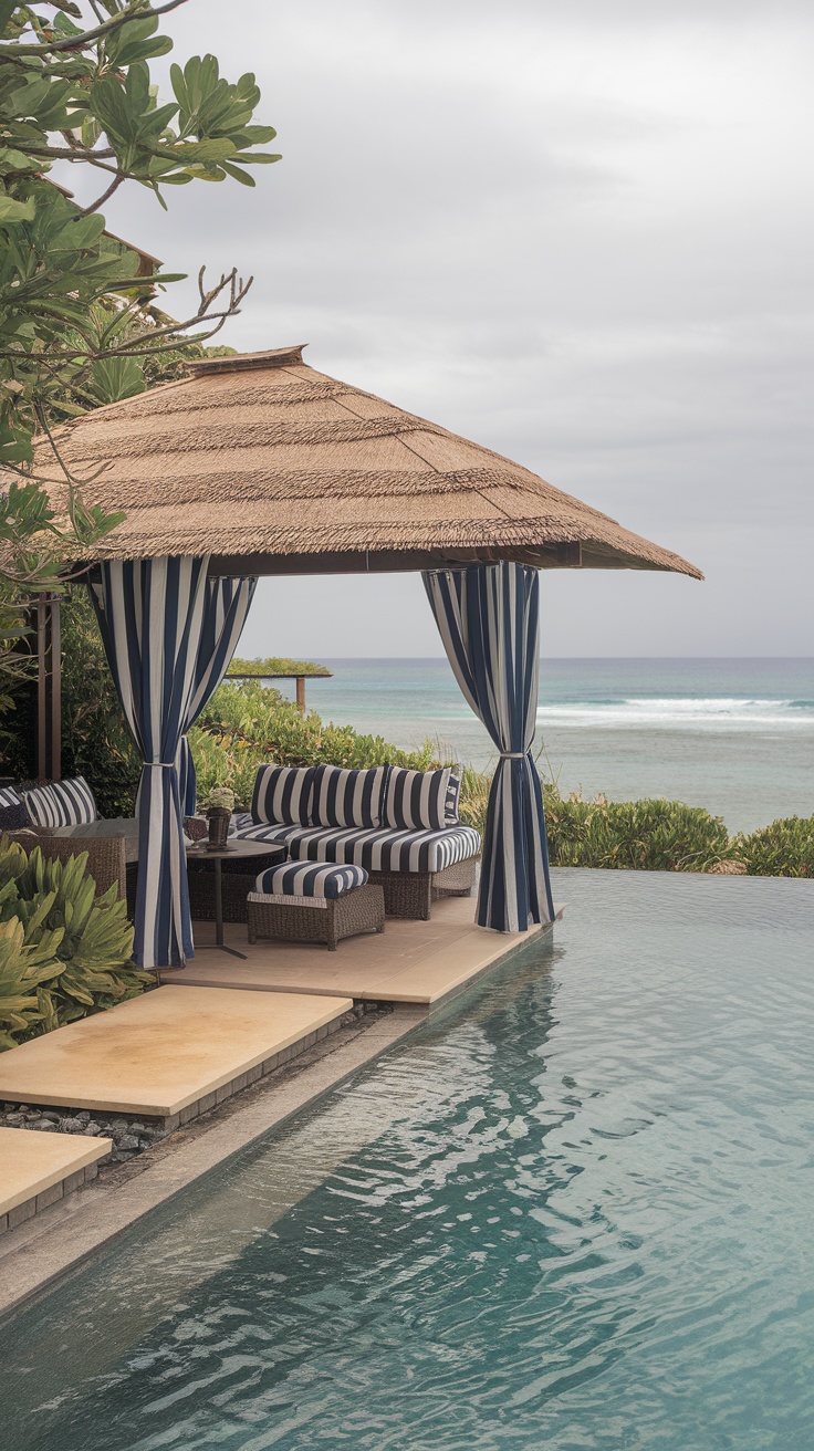 A cozy cabana lounge space by a pool with striped cushions and curtains, overlooking the ocean.